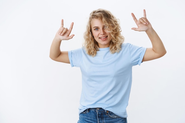 Let roll Cool y elegante, despreocupada, carismática, joven mujer de 20 años con ojos azules en camiseta sonriendo divirtiéndose levantando las manos con un gesto de rock and roll sintiéndose increíble bailando y sonriendo en la fiesta