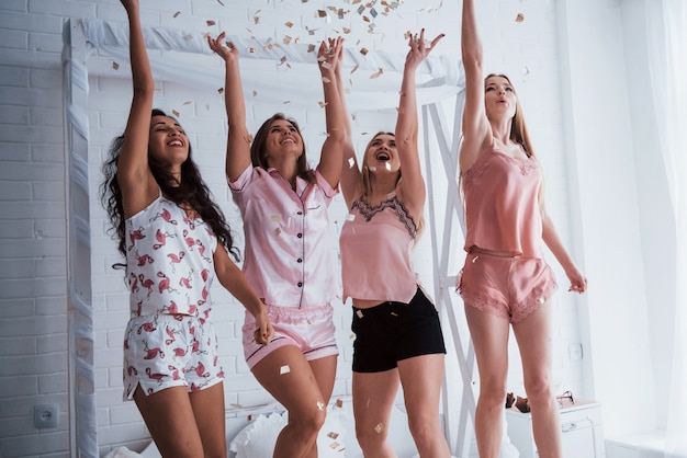 Levanta las manos lo más alto posible Confeti en el aire. Las chicas jóvenes se divierten en la cama blanca en una bonita habitación