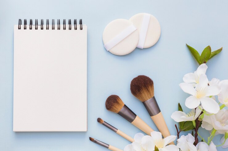 Foto gratuito libreta espiral en blanco; cepillo de esponja y maquillaje con flores blancas sobre fondo azul