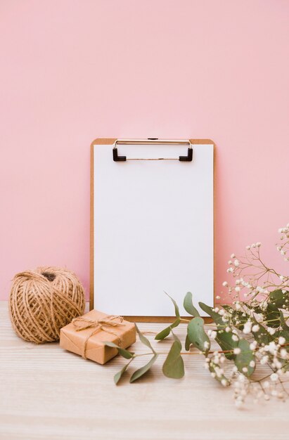 Libro blanco sobre portapapeles con carrete; Caja de regalo y flores de aliento de bebé en el escritorio de madera con fondo rosa