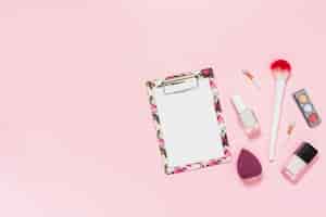 Foto gratuita libro blanco sobre portapapeles con pincel de maquillaje; botella de esmalte de uñas; paleta de sombra de ojos y licuadora sobre fondo rosa