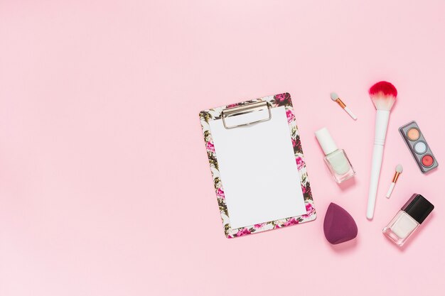 Libro blanco sobre portapapeles con pincel de maquillaje; botella de esmalte de uñas; paleta de sombra de ojos y licuadora sobre fondo rosa