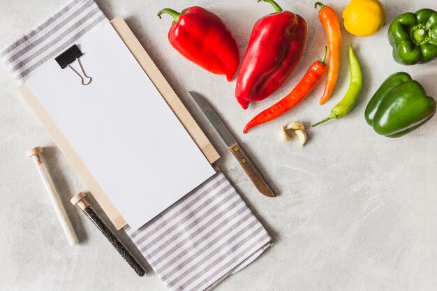 Libro blanco sobre portapapeles con verduras; cuchillo; Tubo de ensayo de sal y pimienta negra.