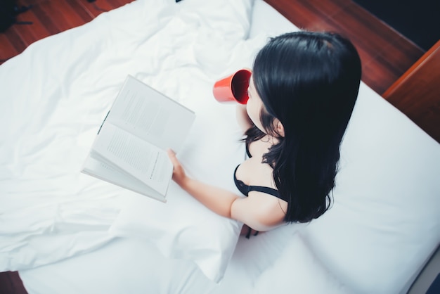 Libro de lectura atractivo de la mujer y sostener la taza de café en cama