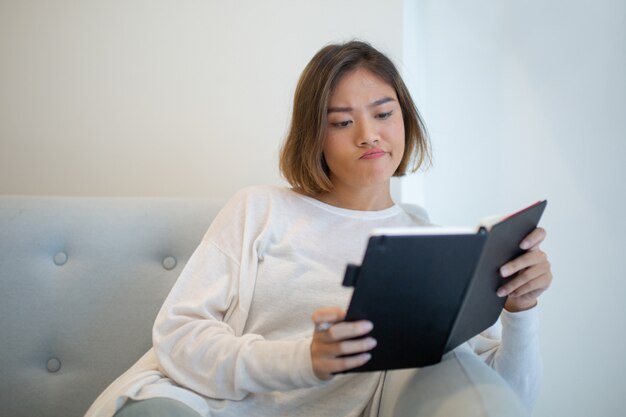 Libro de lectura escéptico de la mujer joven en el sofá en casa