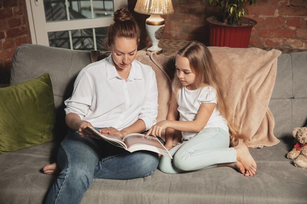 Libro de lectura. Madre e hija durante el autoaislamiento en casa mientras están en cuarentena, tiempo familiar acogedor y confortable, vida doméstica. Modelos sonrientes alegres y felices. Seguridad, prevención, concepto de amor.