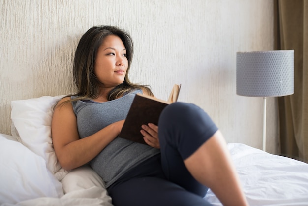 Libro de lectura de mujer japonesa en la cama. Chica en top gris acostado en la cama en posición acogedora con libro impreso