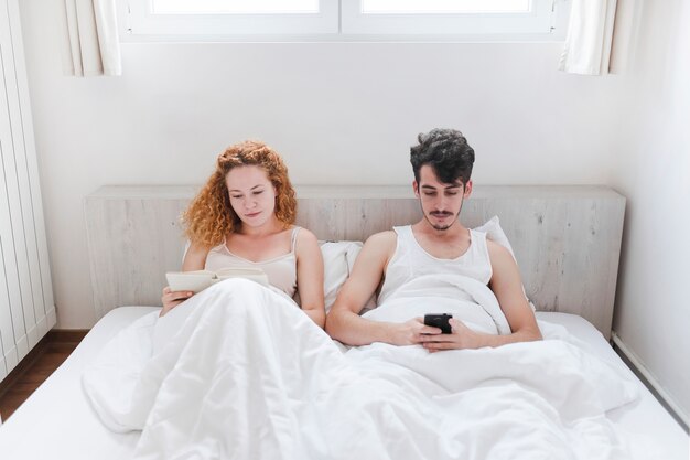 Libro de lectura de la mujer mientras que su marido que usa smartphone en cama