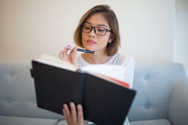 Libro de lectura pensativo de la mujer joven en el sofá en casa