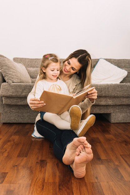 Libro de lectura sonriente de la mujer a la hija cerca del sofá