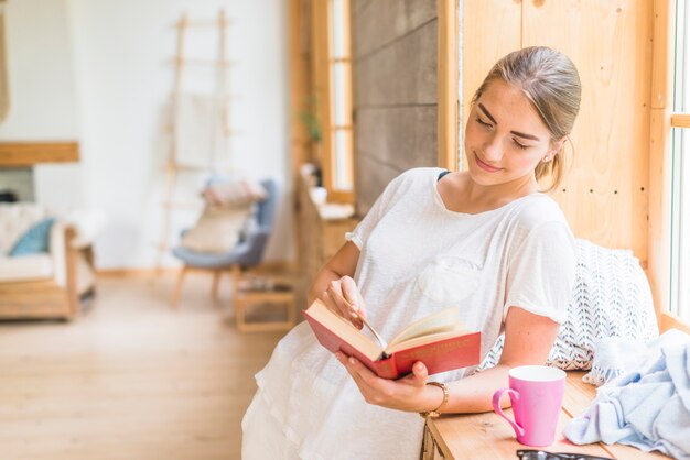 Libro de lectura sonriente de la mujer joven en casa