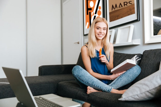 Libro de lectura sonriente de la mujer joven y comer zanahorias en casa