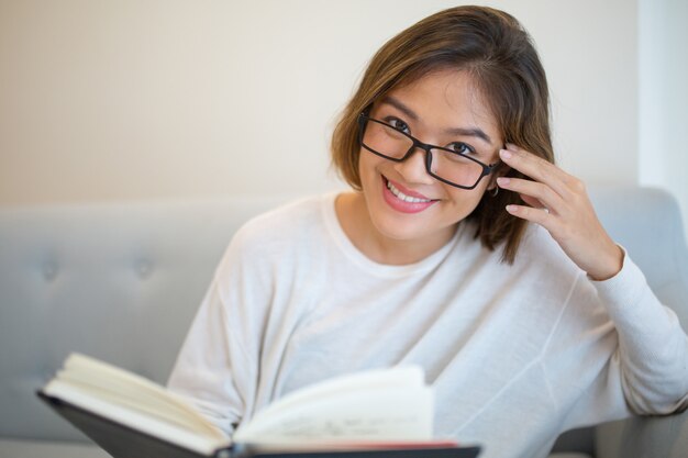 Libro de lectura sonriente de la mujer joven en el sofá en casa