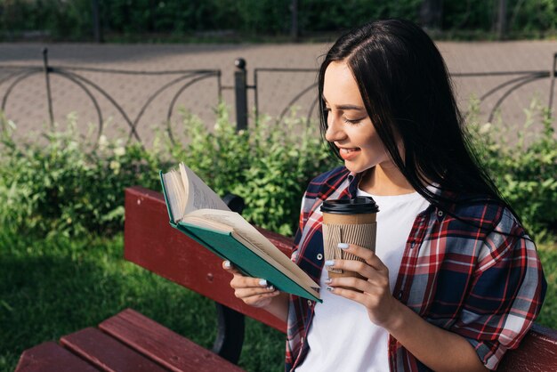 Libro de lectura sonriente de la mujer joven con sostener la taza de café disponible mientras que se sienta en banco en el parque