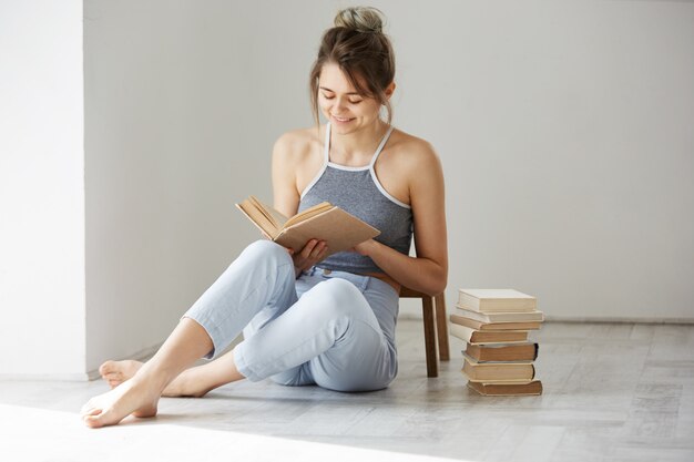 Libro de lectura sonriente de la mujer tierna hermosa joven que se sienta en piso sobre la pared blanca temprano por la mañana.