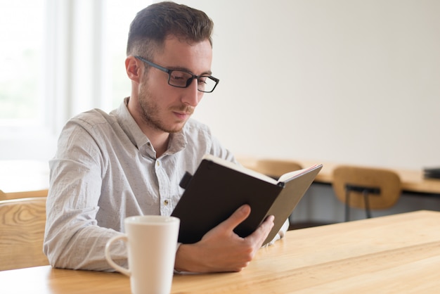 Libro de texto serio de la lectura del estudiante masculino en la tabla en café