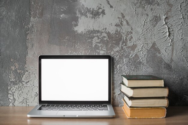 Libros apilados y laptop con pantalla blanca en blanco en superficie de madera