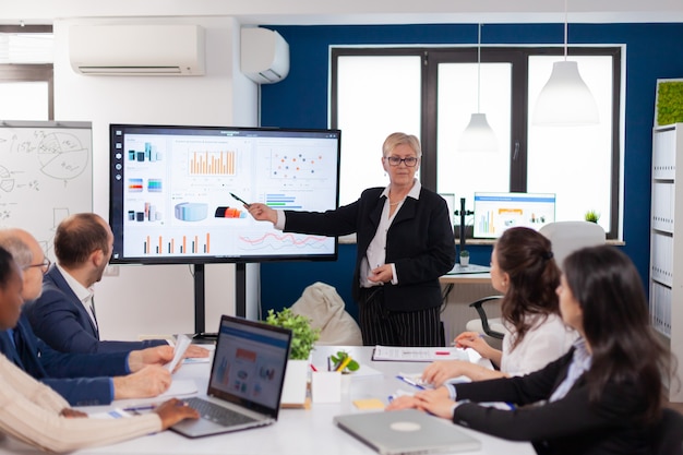 Líder de la empresa senior de intercambio de ideas en la sala de conferencias El personal corporativo discutiendo la nueva aplicación comercial con colegas mirando la pantalla