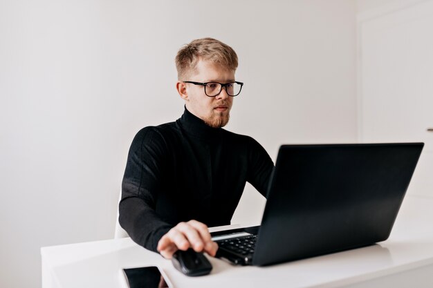 Líder del equipo joven. Hombre joven confiado que trabaja en su escritorio con la computadora portátil y que mira con sonrisa en su oficina ligera.