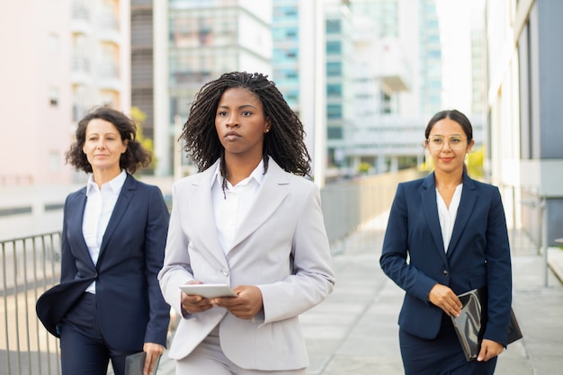 Líder de equipo seguro con tableta durante el paseo. Mujeres empresarias seguras con trajes caminando en la calle. Concepto de trabajo en equipo