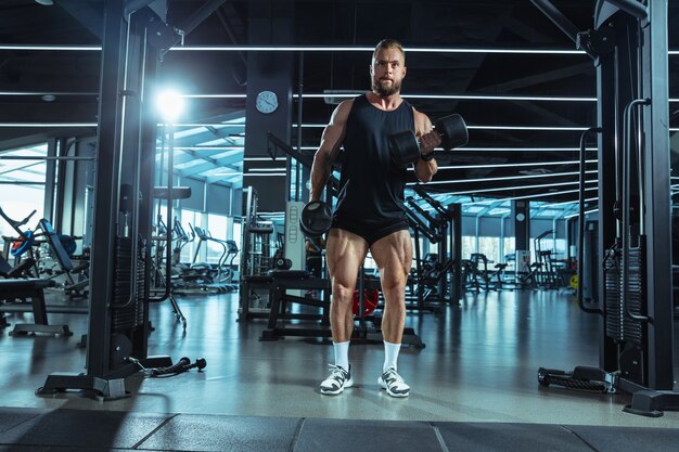 Líder. Joven atleta caucásico muscular entrenando en el gimnasio, haciendo ejercicios de fuerza, practicando, trabajando en la parte superior de su cuerpo con pesas y barra. Fitness, bienestar, concepto de estilo de vida saludable.