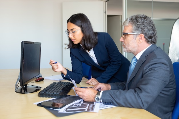 Foto gratuita líder de negocios masculino enfocado y asistente femenina mirando el informe estadístico en el monitor de la pc, sosteniendo gráficos comerciales de papel. vista lateral. concepto de expertos financieros