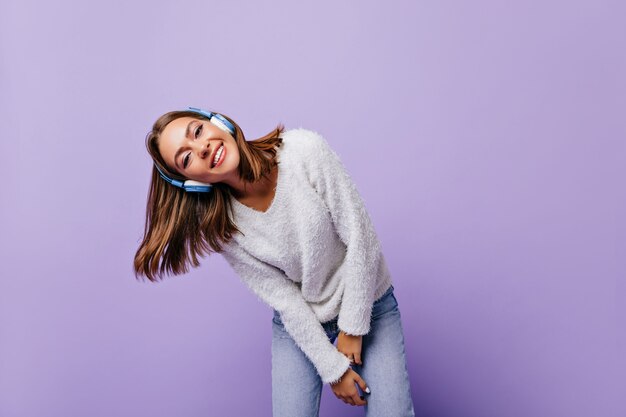 Ligera señorita inclinada, posando relajada y sonriendo amigablemente. Retrato de estudiante en azul auriculares modernos
