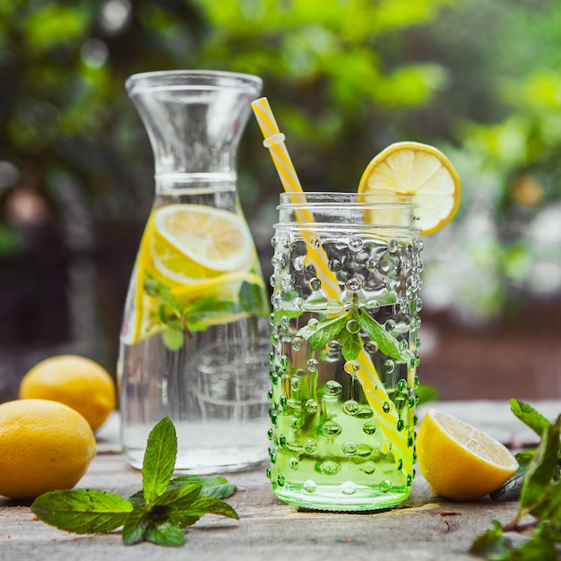 Limonada e ingredientes en jarra de vidrio y tarro en la mesa de madera y jardín, primer plano.