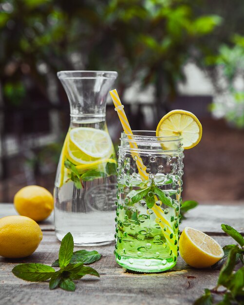 Limonada e ingredientes en jarra de vidrio y tarro en mesa de madera y jardín. vista lateral.