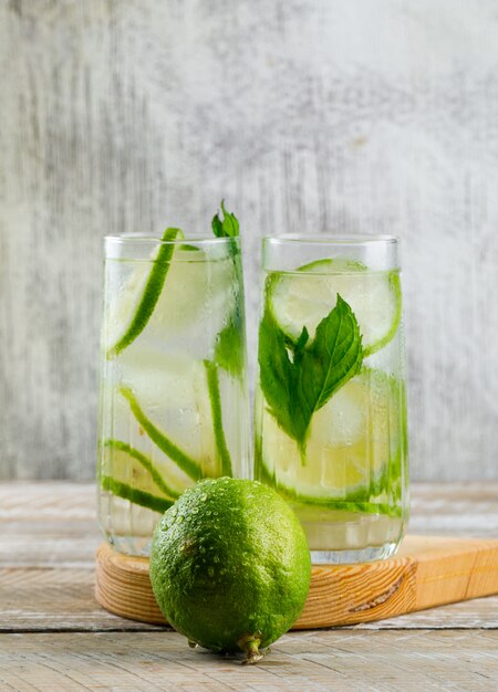 Limonada en vasos con limón, albahaca, vista lateral de tabla de cortar en madera y sucio