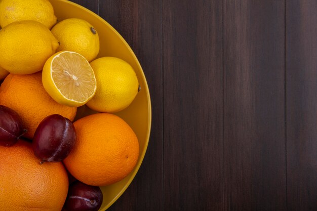 Limones de espacio de copia de vista superior con naranjas, ciruelas y pomelo en un recipiente amarillo sobre un fondo de madera