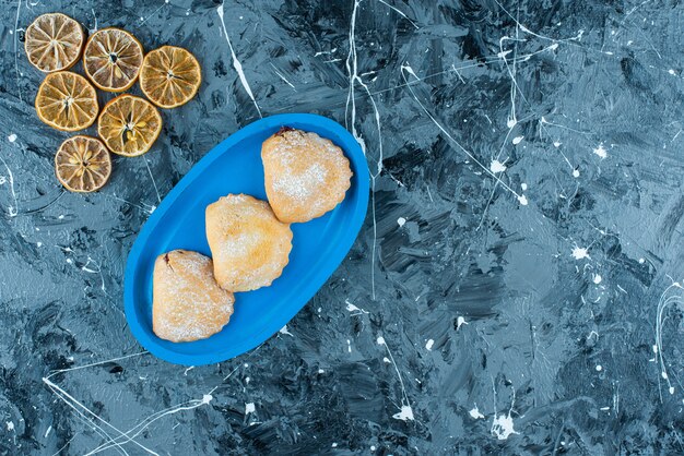 Limones en rodajas junto a tortas en placa de madera, sobre la mesa azul.