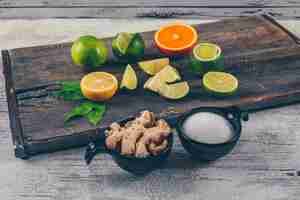 Foto gratuita limones verdes y amarillos y naranja con tazas de sal y vista lateral de jengibre en una bandeja de madera y fondo de madera gris