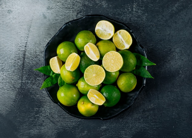 Limones verdes en un plato con rodajas vista superior sobre un fondo negro con textura