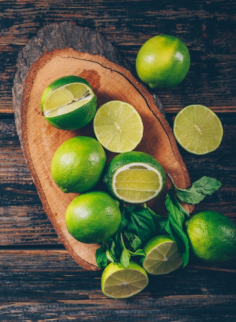 Limones verdes con rodajas y hojas planas sobre una rodaja de madera y fondo de madera oscura.