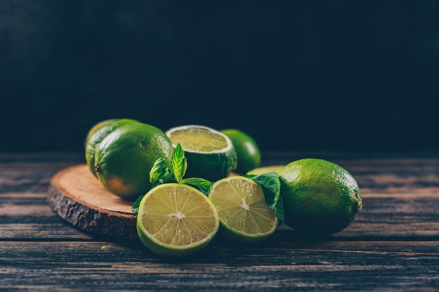 Limones verdes con rodajas y hojas de vista lateral en una rodaja de madera y espacio de fondo de madera oscura para texto