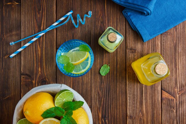 Limones de vista superior en canasta con paño azul, cuchillo de madera y botella de jugo, pajitas sobre superficie de madera.