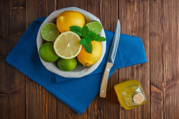 Limones de vista superior en canasta con paño azul, cuchillo de madera y botella de jugo en superficie de madera.