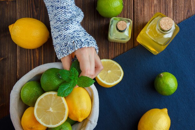 Limones de vista superior en canasta con paño azul, manos sosteniendo hojas sobre superficie de madera. espacio vertical para texto