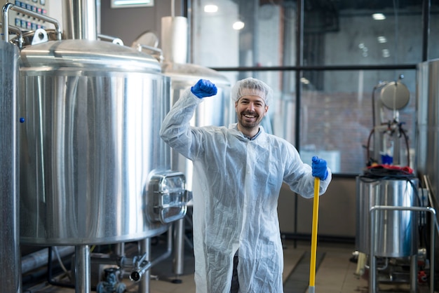 Limpiador industrial profesional en ropa de trabajo de protección sonriendo y sosteniendo el puño en alto en la planta de producción