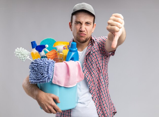 Limpiador joven descontento con gorra sosteniendo el balde con herramientas de limpieza mostrando el pulgar hacia abajo aislado en la pared blanca