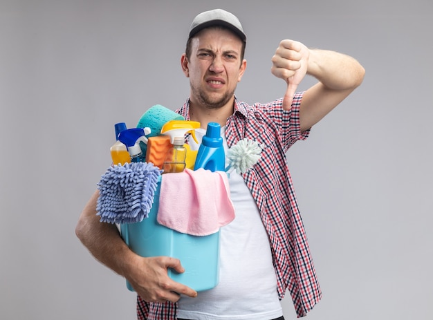 Foto gratuita limpiador joven descontento con gorra sosteniendo un balde con herramientas de limpieza mostrando el pulgar hacia abajo aislado en la pared blanca