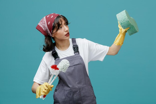 Limpiadora joven concentrada con pañuelo uniforme y guantes de goma sosteniendo cepillos de baño estirando la esponja mirando al costado simulando la limpieza aislada en el fondo azul