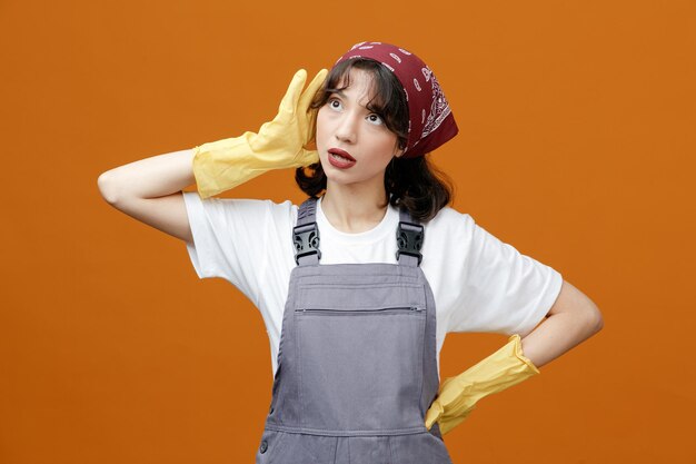 Limpiadora joven impresionada con guantes de goma uniformes y pañuelo manteniendo las manos en la cabeza y en la cintura mirando hacia arriba aislada en el fondo naranja