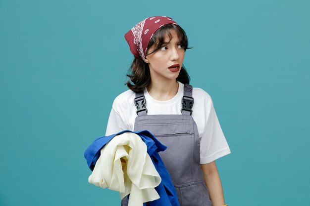 Limpiadora joven impresionada con pañuelo uniforme y guantes de goma sosteniendo ropa sucia mirando al lado aislado en el fondo azul