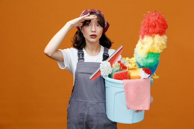Limpiadora joven impresionada con uniforme y pañuelo sosteniendo un cubo de herramientas de limpieza manteniendo la mano en la frente mirando la cámara a distancia aislada en el fondo naranja