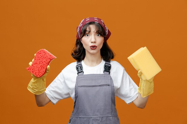 Limpiadora joven impresionada usando guantes de goma uniformes y pañuelo mirando a la cámara mostrando esponjas aisladas en fondo naranja