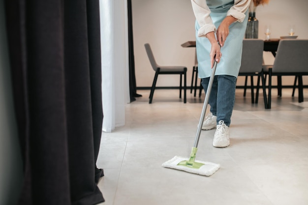 Foto gratuita limpieza de la cocina. una mujer joven en delantal limpiando el piso en la cocina