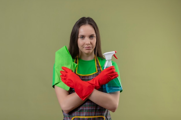 Foto gratuita limpieza joven vistiendo uniforme en guantes rojos sosteniendo spray de limpieza con trapo cruzando las manos sobre la pared verde aislada