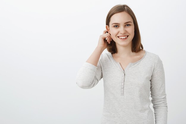 Linda adolescente emotiva en blusa casual moviendo el cabello detrás de la oreja aleteada y sonriendo ampliamente sintiéndose tímida y alegre al ser invitada a jugar con una compañía interesante sobre una pared blanca